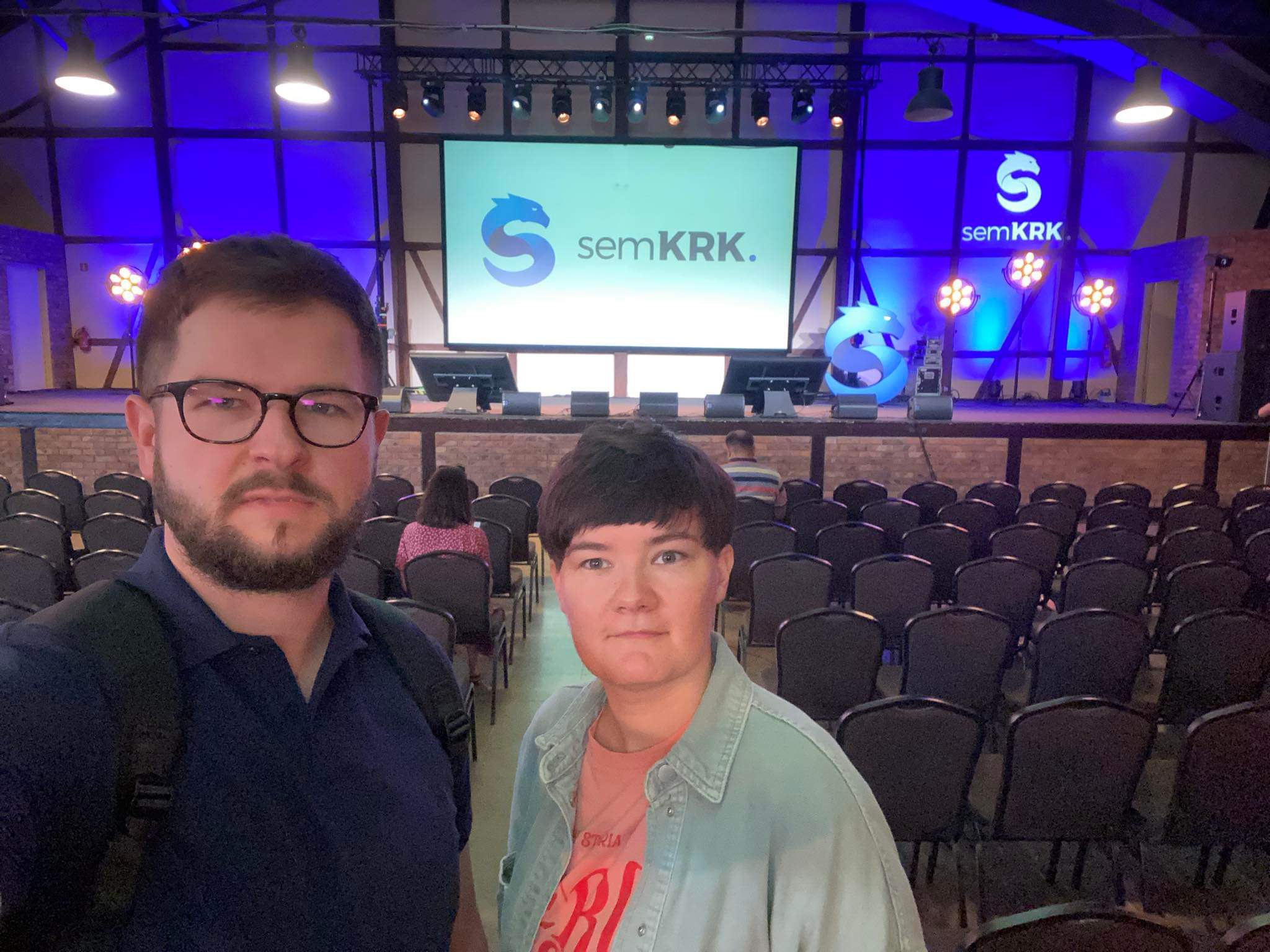 Sala konferencyjna z dwoma osobami robiącymi sobie selfie na tle sceny z logo semKRK i dużą liczbą krzeseł ustawionych przed sceną.